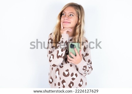Thinking dreaming caucasian teen girl wearing animal print sweater over white background using mobile phone and holding hand on face. Taking decisions and social media concept.