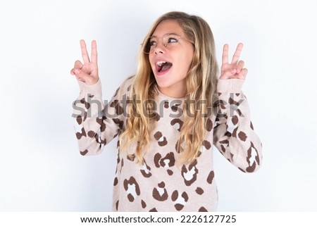 Isolated shot of cheerful caucasian teen girl wearing animal print sweater over white background makes peace or victory sign with both hands, feels cool.