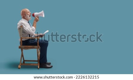 Confident professional film director sitting on the director's chair and shouting with a megaphone, film industry concept Royalty-Free Stock Photo #2226132215