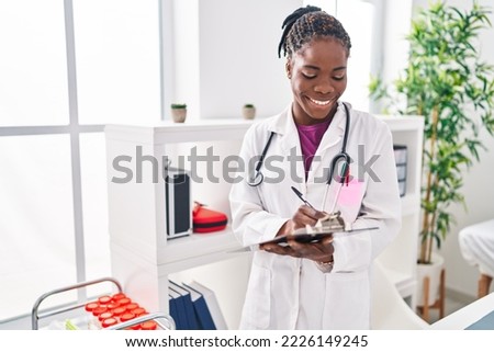 African american woman wearing doctor uniform writing on clipboard at clinic