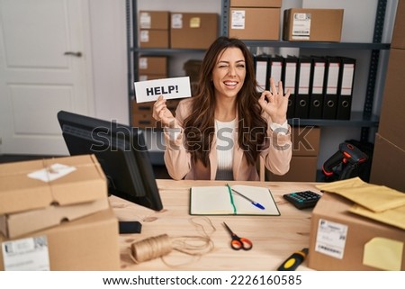 Young brunette woman working at small business ecommerce holding help banner doing ok sign with fingers, smiling friendly gesturing excellent symbol 