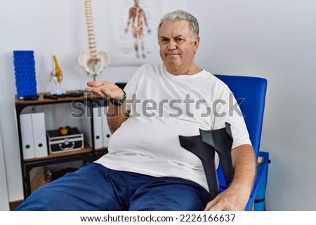 Senior caucasian man at physiotherapy clinic holding crutches pointing aside with hands open palms showing copy space, presenting advertisement smiling excited happy 