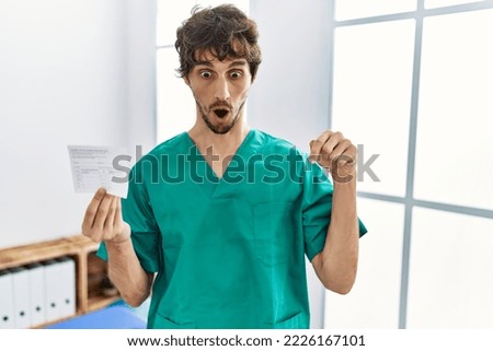 Young hispanic doctor man holding covid record card pointing down with fingers showing advertisement, surprised face and open mouth 