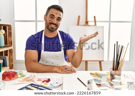 Young hispanic man with beard at art studio with painted face smiling cheerful presenting and pointing with palm of hand looking at the camera. 