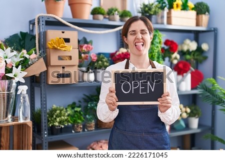 Brunette woman working at florist holding open sign sticking tongue out happy with funny expression. 