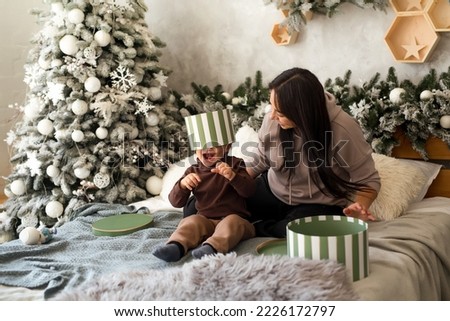 Mother with infant child having fun at Christmas with gift boxes. Baby boy excited about festive surprise and play at home. Family celebration and preparation for New Year