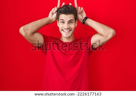Young hispanic man standing over red background posing funny and crazy with fingers on head as bunny ears, smiling cheerful 