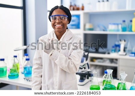 African american woman working at scientist laboratory smiling cheerful pointing with hand and finger up to the side 