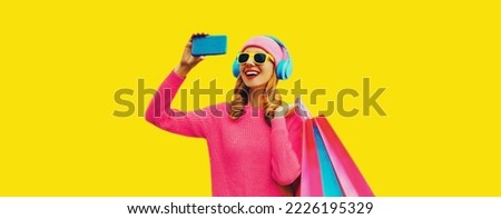 Stylish happy smiling woman taking selfie with phone in headphones with shopping bags wearing knitted sweater, pink hat on yellow background