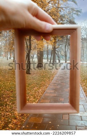 The hand holds a wooden frame in which the autumn city alley is visible on a cloudy rainy day. Copy space