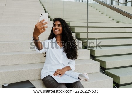 Young indian woman taking selfie photo on cellphone while sitting on stairs outdoors
