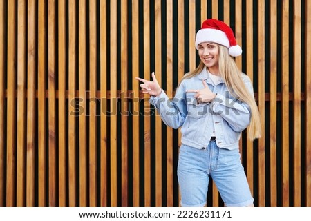 New year concept, young smiling caucasian woman in christmas santa hat jeans outfit points on blank copy space in front wood wall background outdoor