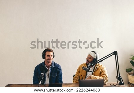 Young male podcaster interviewing a guest on his show. Vibrant young man co-hosting an audio broadcast with a guest. Two college content creators recording a live show in a studio.