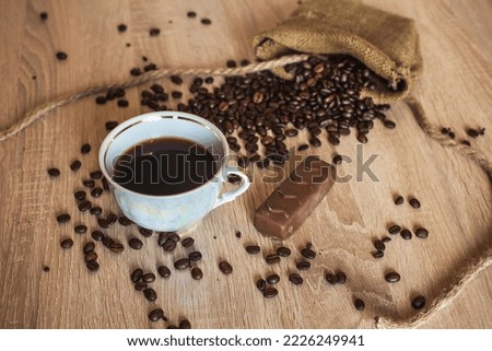 Roasted coffee beans in little bag with ropes and cup of coffee with drink and chocolate on brown wooden surface, in the morning for breakfast. Image for your creativity, design or illustrations.