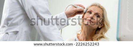 blonde woman looking away near doctor doing ultrasound examination of her head, banner Royalty-Free Stock Photo #2226252243