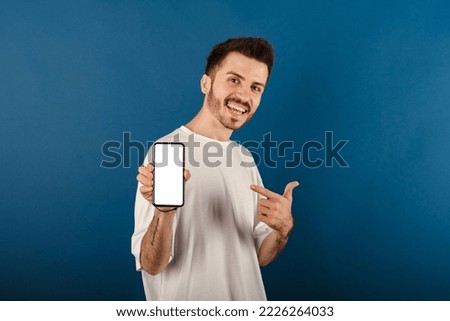 Cheerful caucasian man wearing casual clothes posing isolated over blue background showing to camera and pointing at device. Gadget with empty free space for mock up. White blank screen.