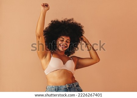 Beautiful woman with hair dancing cheerfully while wearing a bra and jeans. Happy plus size woman celebrating her natural body and curly hair. Body positive young woman having fun in a studio. Royalty-Free Stock Photo #2226267401