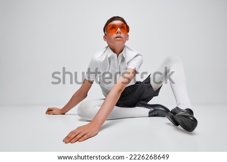 Portrait of stylish boy posing in black and white clothes with red glasses isolated over grey background. Modelling . Concept of modern fashion, art photography, style, queer, uniqueness, ad