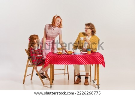 Portrait of beautiful woman, man and little girl sitting at the table and having breakfast isolated over grey background. Concept of beauty, retro style, fashion, 60s, 70s, family. Copy space for ad Royalty-Free Stock Photo #2226268775