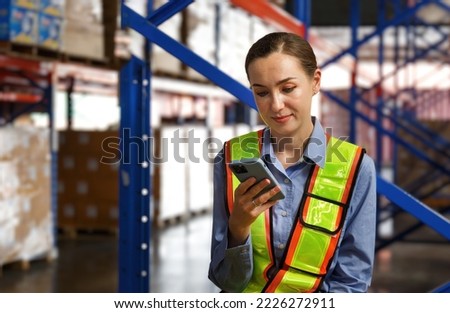 Female warehouse engineer wearing hard hat checking goods in warehouse and controlling load from cargo ship to import and export by reporting on the smartphone.