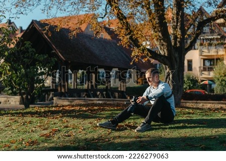 Professional Photographer Taking Picture Of Beautiful Autumn Park. Man Professional Photographer Sit With Camera And With Smartphone In Autumn Park. Retouched, Vibrant Colors, Brownish Tones.