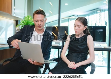 Two young Asian business woman discussing project and report in laptop and digital tablet while standing and analyzing data on statistics and brainstorming information in modern office