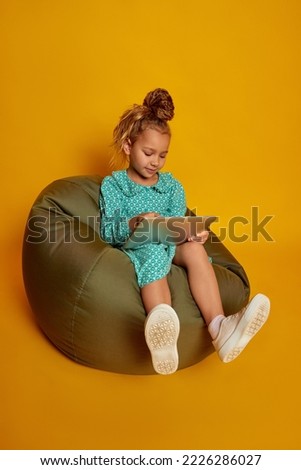 Portrait of nice pretty cheerful wavy-haired girl sitting and using laptop isolated on yellow studio color background. Concept of children positive emotions, beauty, facial expressions. Copy space, ad