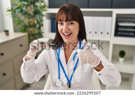 Young brunette doctor woman holding syringe smiling happy and positive, thumb up doing excellent and approval sign 