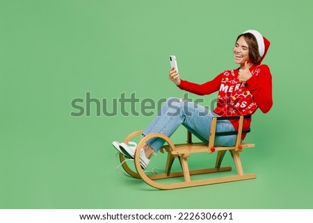 Full length merry young woman wear xmas sweater Santa hat posing sit on sleigh sledding do selfie shot on mobile cell phone isolated on plain pastel light green background Happy New Year 2023 concept