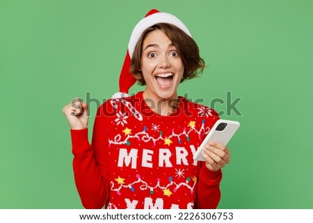 Merry young woman wears knitted xmas sweater Santa hat posing hold in hand use mobile cell phone do winner gesture isolated on plain pastel light green background. Happy New Year 2023 holiday concept Royalty-Free Stock Photo #2226306753