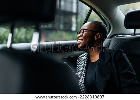 A black ethnicity businesswoman is in her car sitting on the backseat using her mobile phone and smiling. Royalty-Free Stock Photo #2226310807