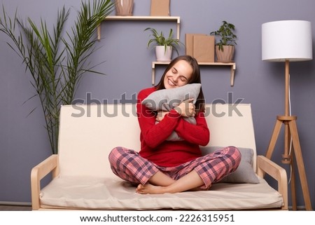 Image of pretty happy beautiful young adult woman wearing red jumper and pajama pants embracing pillow, being in good mood, relaxing at weekend, rejoicing her rest, sitting on sofa in home interior.