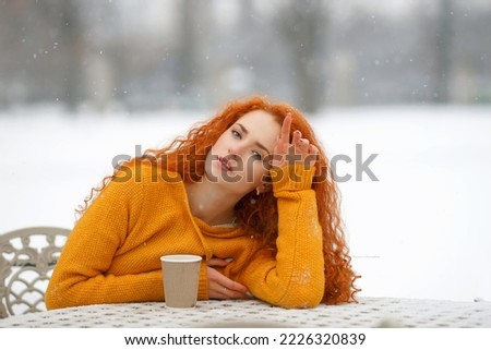                Close-up portrait of a red-haired beautiful girl looking at the camera sitting at the table on the street in winter, it's snowing hot with coffee Cold winter,
 white snowflake 