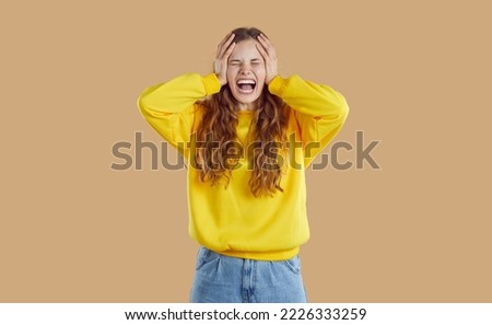 Negative human emotions. Emotional teenage girl because of anger and resentment screams and cries on beige background. Girl grabs her head with her hands. Concept of hatred, rage and frustration. Royalty-Free Stock Photo #2226333259
