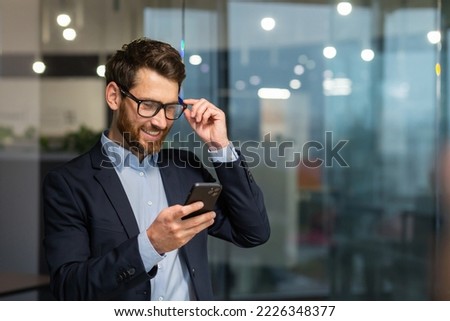 Successful financier investor works inside office at work, businessman in business suit uses telephone near window, man smiles and reads good news online from smartphone. Royalty-Free Stock Photo #2226348377