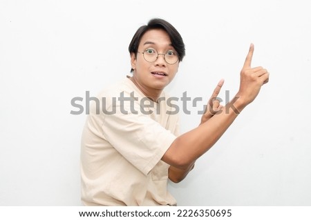asian young man in casual beige tshirt pointing finger showing something good. man with glasses shocked happily pointing up with copy space isolated over white background. advertisment model concept.
