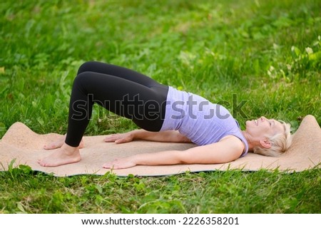 Athletic attractive middle aged woman practicing yoga doing yoga in nature in bridge pose. Glute bridge pose.The concept of stretching, pilates, doing sports in nature. Royalty-Free Stock Photo #2226358201