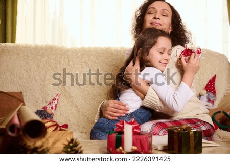 Charming little girl, beloved daughter gently leans on the shoulders of her mother hugging her, receiving a cute Christmas present. Beautiful gifts on the table in foreground. Happy carefree childhood