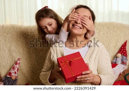 Pretty loving mother smiling a cheerful toothy smile, while her lovely daughter covers her eyes with her hands, offers her a Christmas present in stylish red box, sitting together on sofa at home
