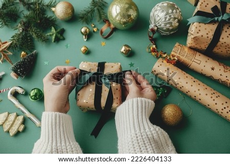 Merry Christmas! Hands wrapping stylish christmas gift, golden wrapping paper, green ribbon and festive decorations on green background. Wrapping christmas gifts concept. Happy Holidays Royalty-Free Stock Photo #2226404133