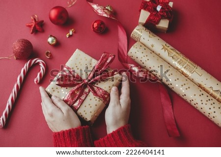Merry Christmas! Hands holding stylish christmas gift, golden wrapping paper, ribbon and festive decorations on red background. Seasons greetings. Happy Holidays