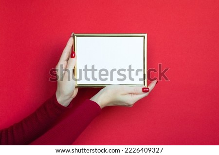 The hands of a white woman in red holding horizontal golden frame over a red background