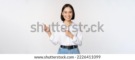 Image of smiling young office lady, asian business entrepreneur pointing fingers left, showing client info, chart of banner aside on copy space, white background. Royalty-Free Stock Photo #2226411099