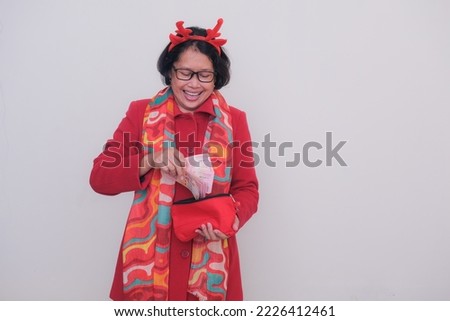 A middle-aged Asian woman wearing a red coat, Christmas-themed bandana and colorful shawl keeps some money in a red clutch; smiling, happy expression.