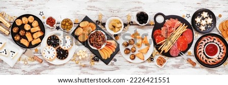 New Years Eve theme appetizer table scene. Above view on a white wood banner background. Charcuterie board, champagne, clock cookies, tuxedo brie cheese and a variety of party food.