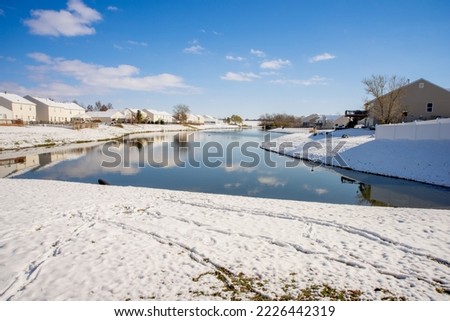 Snow around a lake in winter. Houses line the shore. Footprints can be seen in the snow. The sky is bright blue with puffy clouds. The water is a dark blue green. Royalty-Free Stock Photo #2226442319