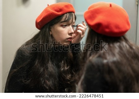 Young elegant girl in a red beret doing her makeup with eyeliner. Beautiful female wearing black dress and red beret, looking in a mirror.