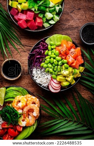 Hawaiian poke bowl set: tuna, salmon, shrimp with avocado, mango, radish, rice and other ingredients. Soy sauce and sesame dressing. Wooden table background, palm leaves, top view Royalty-Free Stock Photo #2226488535