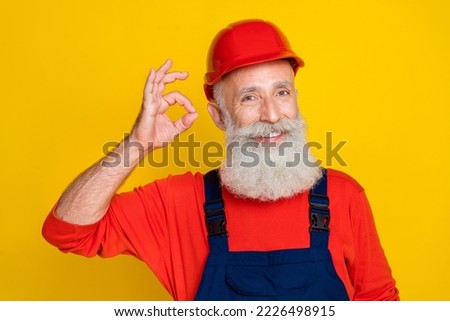 Photo of pretty cheerful senior guy dressed uniform overall red hardhat showing okey sign isolated yellow color background