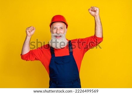 Photo of cheerful lucky age man workwear overall red hard hat smiling rising fists isolated yellow color background